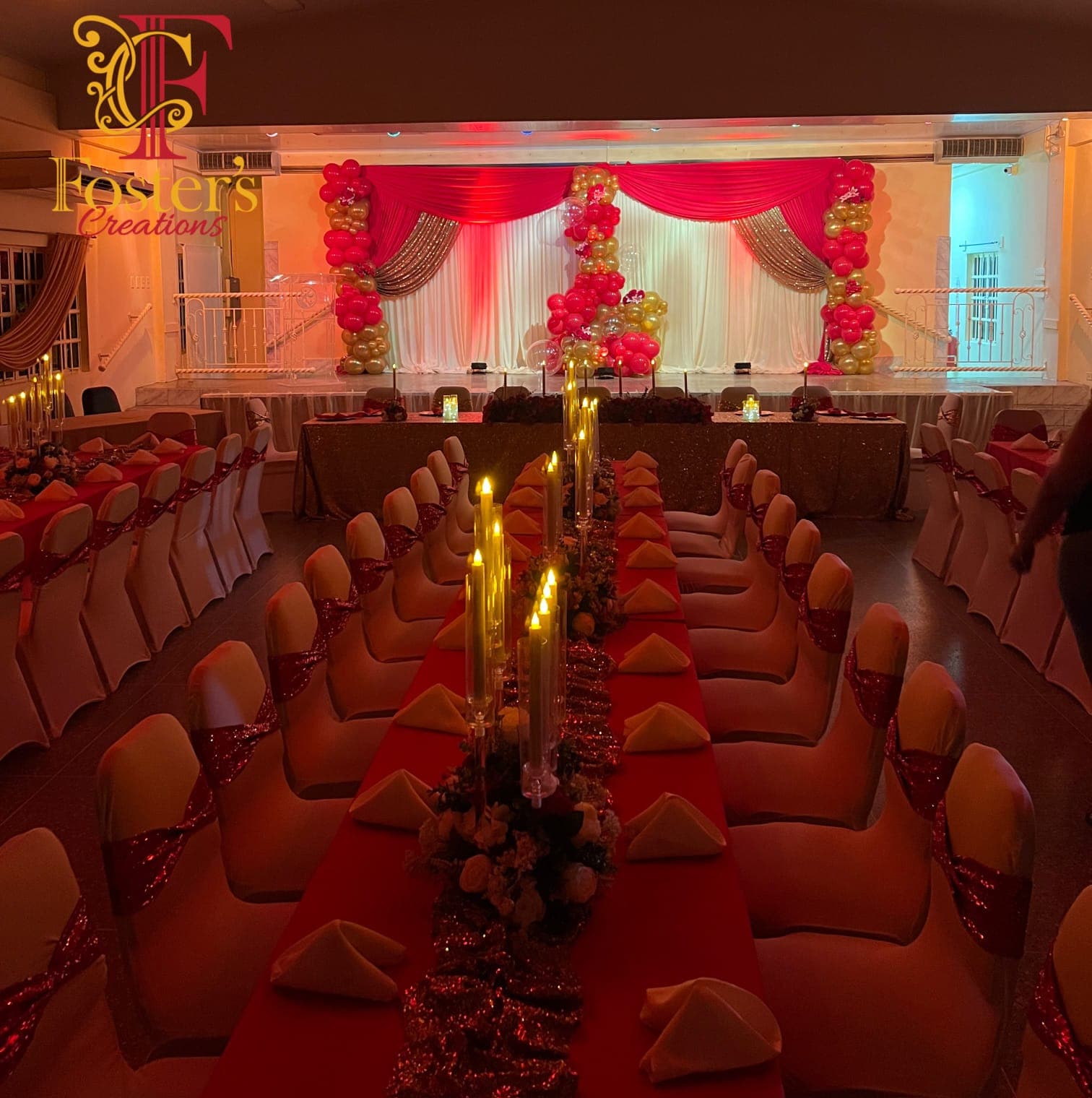 Hall reception under warm uplighting: a crimson, gold and ivory swagged stage backdrop framed by red and gold balloon columns, fronted by a long guest table in red linen with a sequin runner, taper candles and white chair covers tied with red sequin sashes