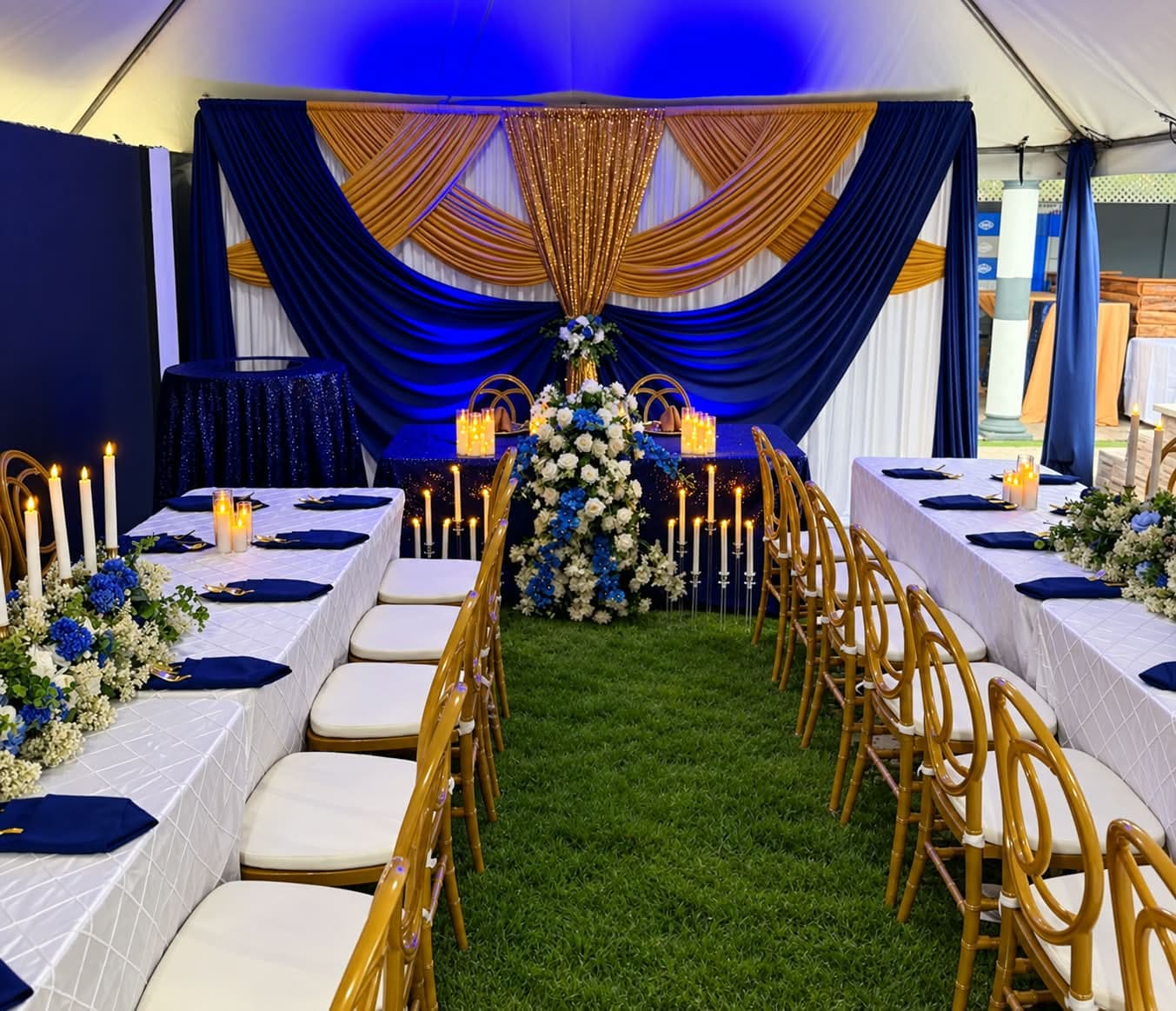 Marquee reception on grass: a navy, gold and ivory swagged backdrop centred on a shimmering gold fairy-light curtain, with a navy sequin head table, gold chiavari chairs, white linen guest tables in navy napkins and a tall blue and white floral column
