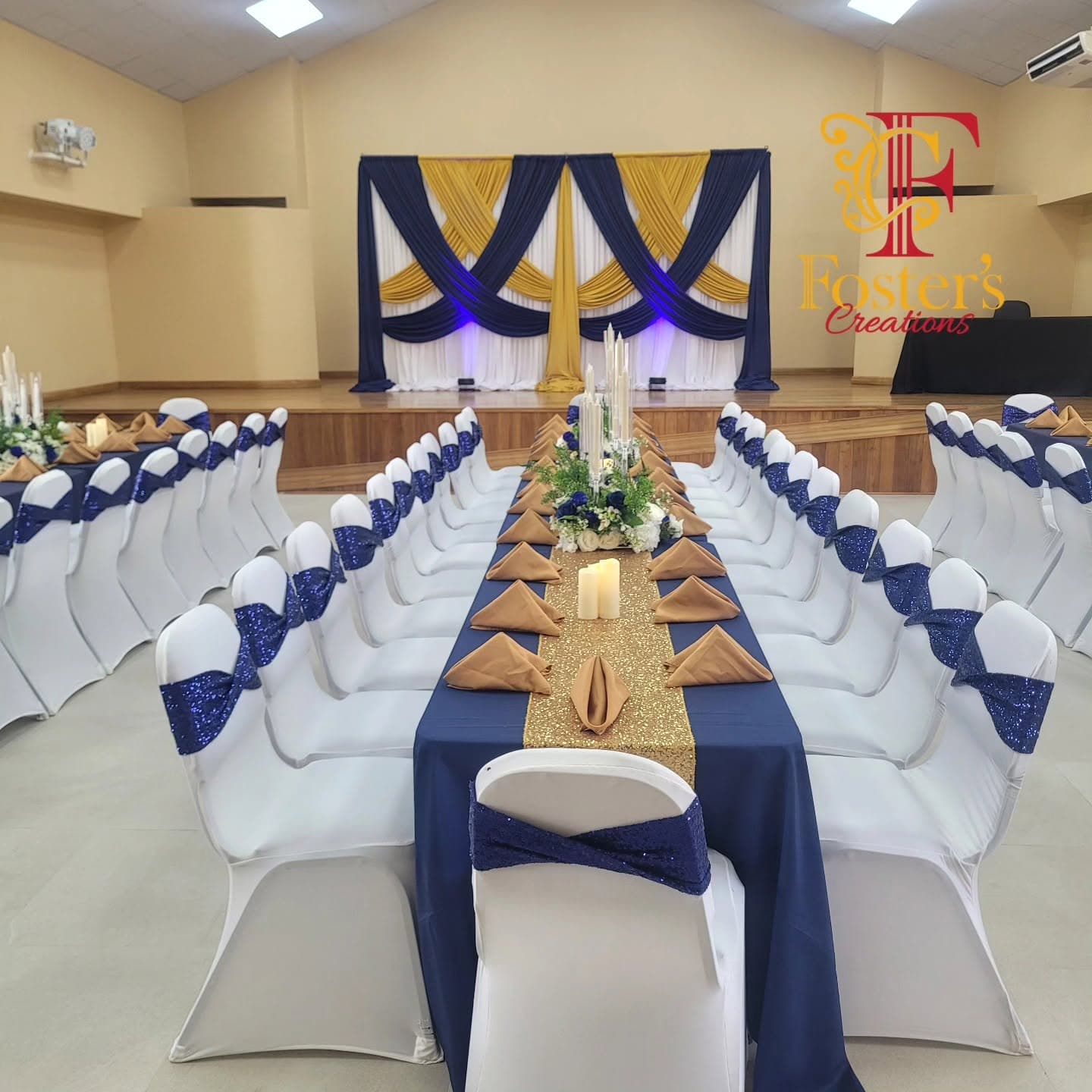 Hall reception with a navy, gold and white draped stage backdrop, long head table in navy linen with a gold sequin runner and white chair covers tied with navy sashes